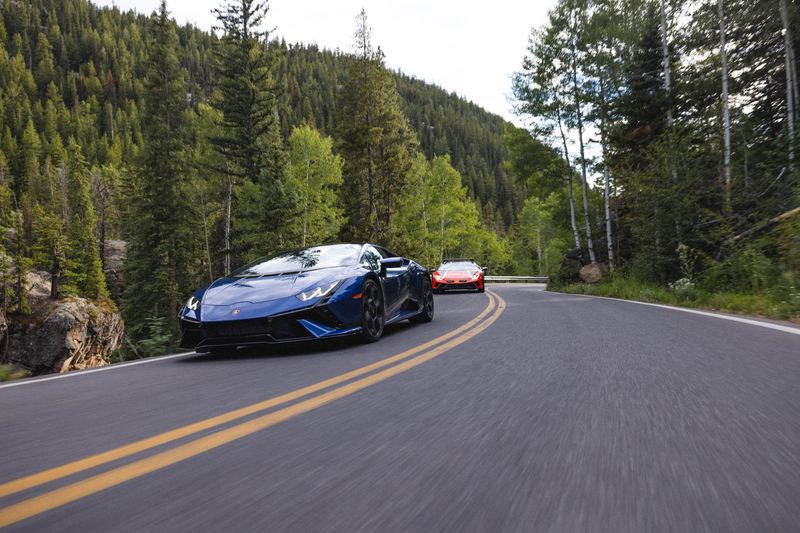 A blue Lamborghini on a forest mountain road with a second car behind
