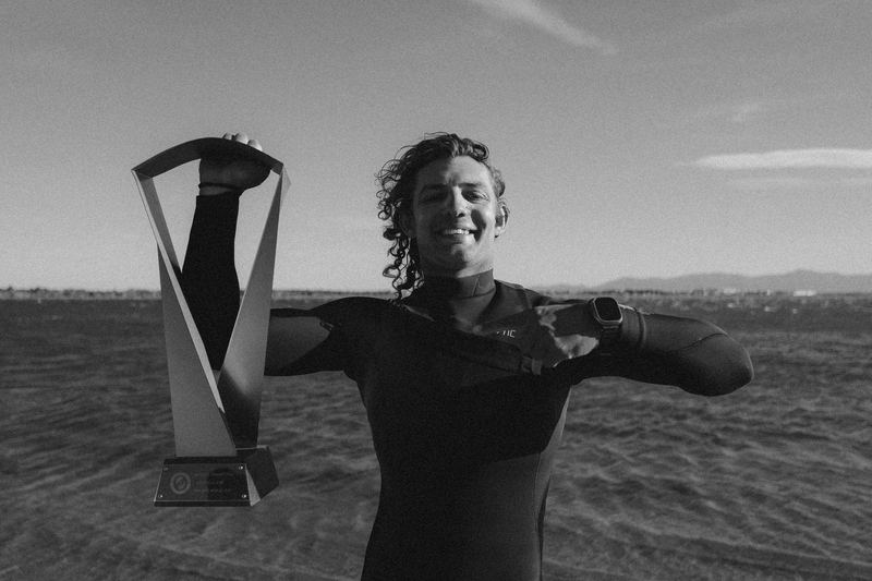 A kitesurfer holding up a competition trophy on the beach