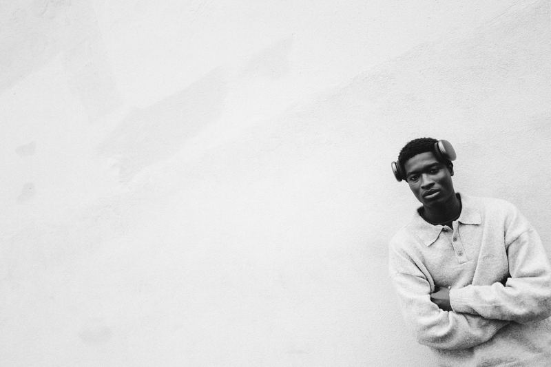 A young man leaning against a white wall, arms crossed